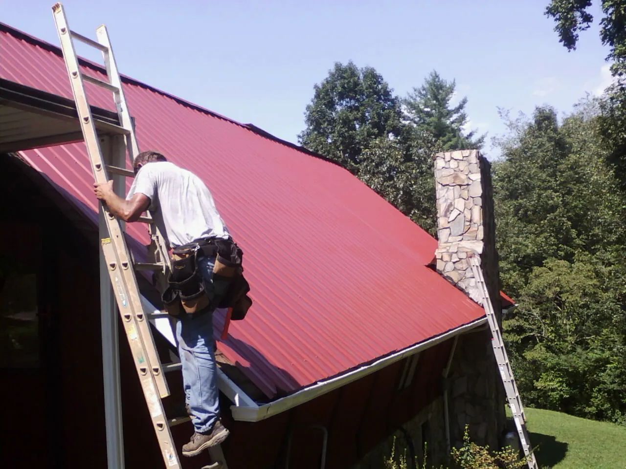 Roofer on a ladder beside a red metal roof and stone chimney — Soffit & Fascia Repair by Roofing Brothers in Los Banos